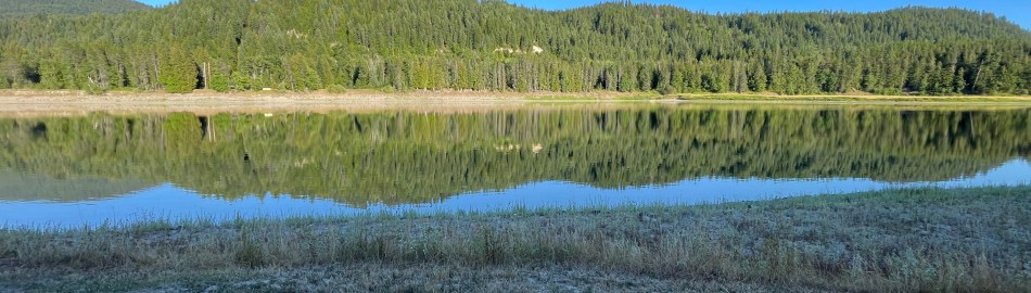 Tree filled range reflected in calm river in bright sunshine.